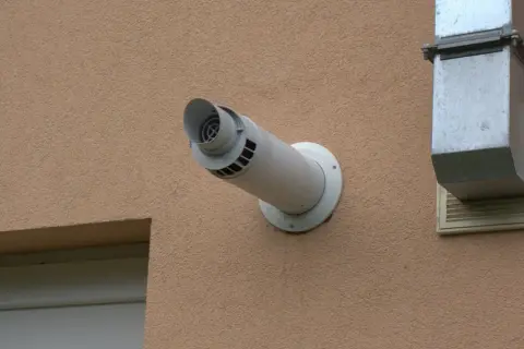 Odprowadzenie spalin z pieca gazowego przez ścianę, zgodnie z przepisami, wymaga odpowiedniej instalacji. Widoczny biały kominek wentylacyjny i metalowy kanał.