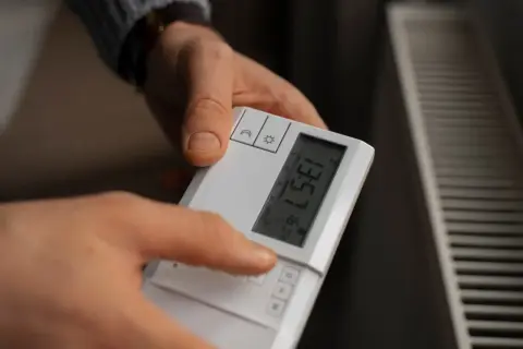 Dłoń naciska przycisk na białym sterowniku, pokazując jak ustawić temperaturę na sterowniku. Obok widać grzejnik.