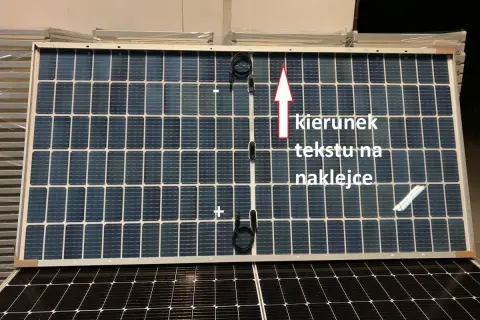 Panel fotowoltaiczny z oznaczeniami plus i minus. Strzałka wskazuje kierunek tekstu na naklejce.