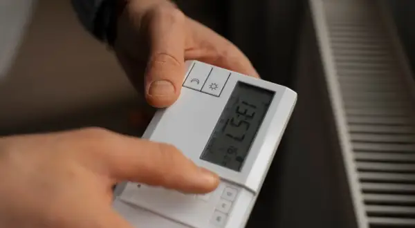 Dłoń naciska przycisk na białym sterowniku, pokazując jak ustawić temperaturę na sterowniku. Obok widać grzejnik.