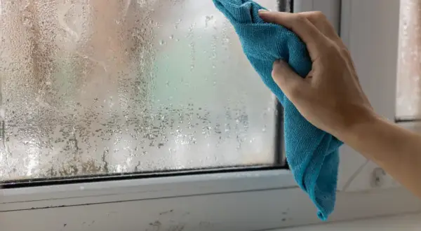 Ręka z niebieską ściereczką wyciera zaparowane okno. Skraplająca się woda na szybie to znak, że trzeba pomyśleć, jak pozbyć się wilgoci w pokoju.