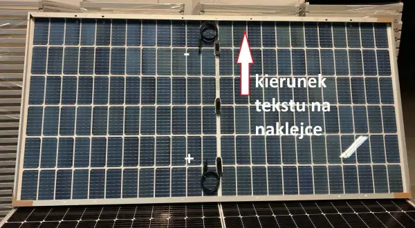 Panel fotowoltaiczny z oznaczeniami plus i minus. Strzałka wskazuje kierunek tekstu na naklejce.