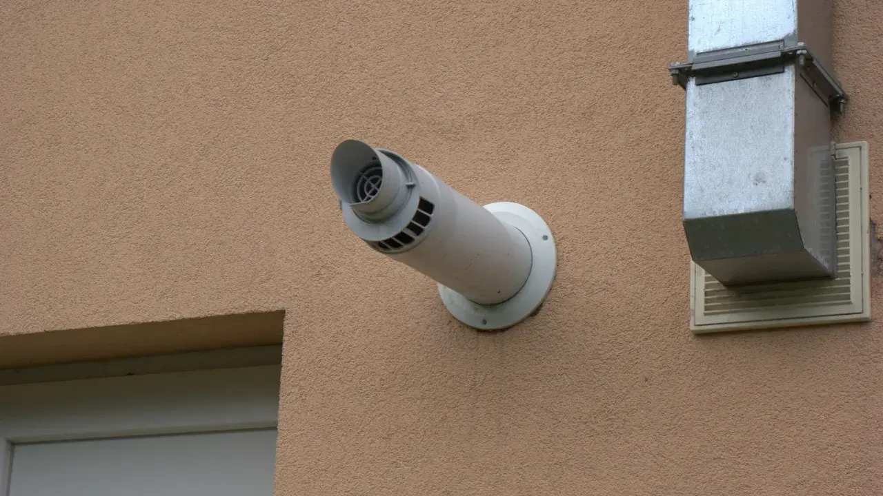 Odprowadzenie spalin z pieca gazowego przez ścianę, zgodnie z przepisami, wymaga odpowiedniej instalacji. Widoczny biały kominek wentylacyjny i metalowy kanał.