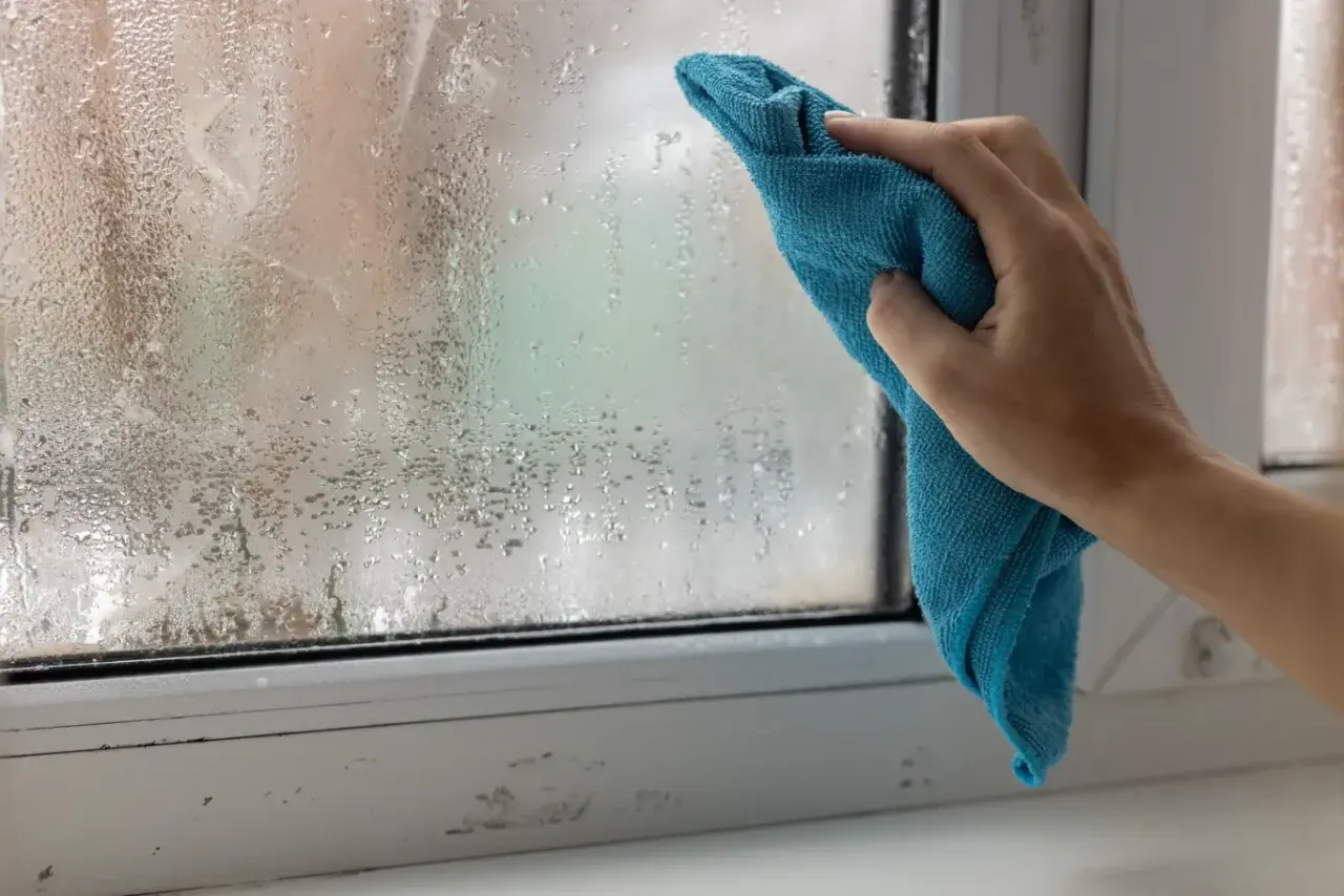 Ręka z niebieską ściereczką wyciera zaparowane okno. Skraplająca się woda na szybie to znak, że trzeba pomyśleć, jak pozbyć się wilgoci w pokoju.
