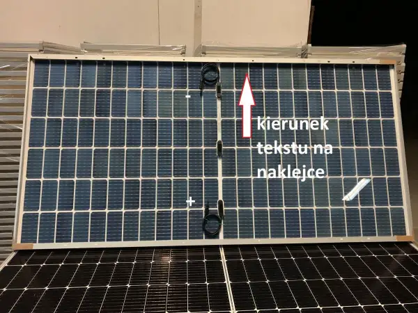 Panel fotowoltaiczny z oznaczeniami plus i minus. Strzałka wskazuje kierunek tekstu na naklejce.