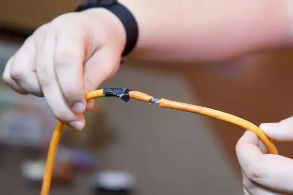 Ręce trzymające przerwany pomarańczowy kabel, pokazując, jak połączyć kable bez lutowania za pomocą taśmy izolacyjnej.